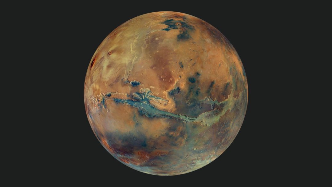 Imagen compuesta por un mosaico de imágenes que muestra el globo terráqueo de Marte sobre un fondo oscuro. El disco del planeta presenta manchas amarillas, naranjas, azules y verdes, todas ellas con un tono gris apagado, que representa la composición variable de la superficie. Crédito: ESA/DLR/FU Berlin/G. Michael, CC BY-SA 3.0 IGO